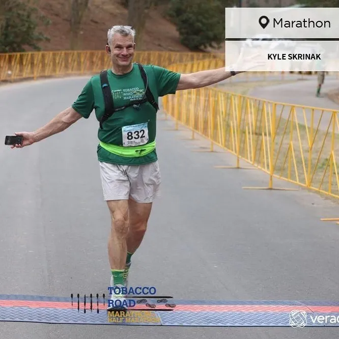 Kyle crossing the Tobacco Road Marathon finish line with arms wide, phone in hand, wearing bib 832, green shirt and socks