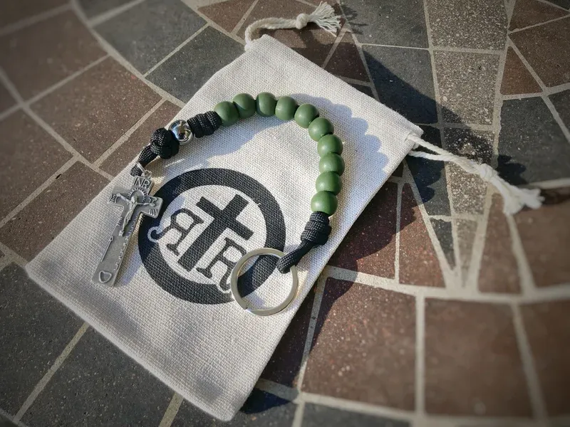 Green-beaded pocket rosary with silver crucifix and keyring, resting on a canvas drawstring bag