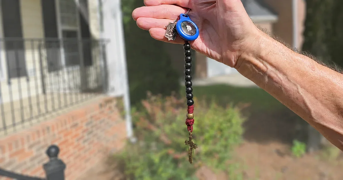 A hand holding a black-beaded pocket rosary with a blue Apple AirTag attached